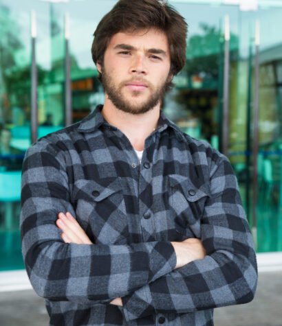 Serious young man standing outdoors with his arms crossed. Handsome guy wearing casual shirt, looking at camera and standing with glass wall in background. Young man portrait concept. Front view.