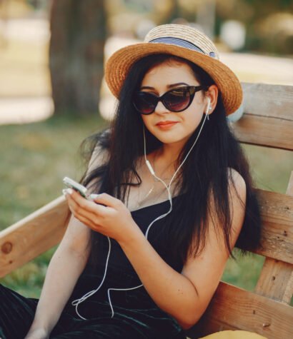 Beautiful and stylish girl in hats sitting in a summer park on pallets with phone and headphones